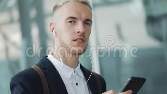 年轻迷人的商人站在商务中心附近带着智能手机和耳机他看着摄像机视频的预览图