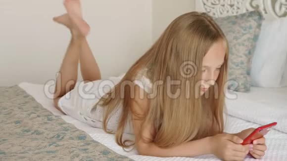 少女躺在白床上看手机年轻女孩在白色卧室的床上用智能手机放松女孩视频的预览图