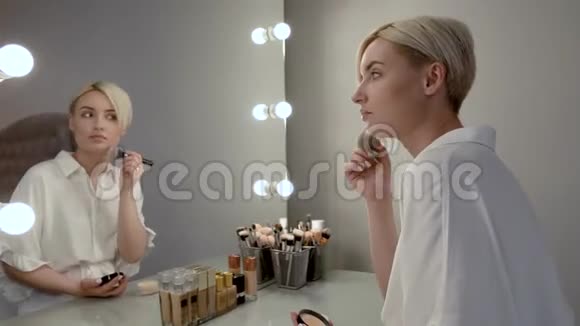 年轻的金发女孩短发坐在一面大镜子前用大圆底粉涂在她脸上视频的预览图