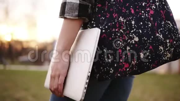 从穿牛仔裤的女孩的侧面看她手里拿着一个彩色的袋子和一台笔记本电脑穿过公园关门视频的预览图