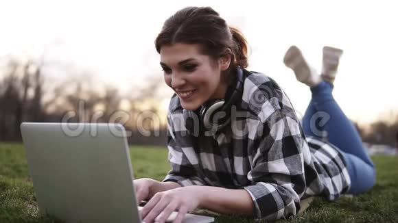 美丽的女孩躺在公园的草地上用笔记本电脑聊天脖子上的耳机她玩得很开心视频的预览图