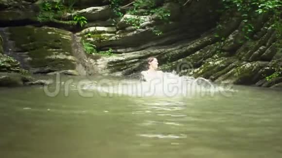 在绿色热带森林中瀑布附近的小山湖里年轻的美女双手泼水视频的预览图