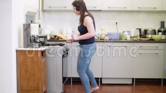 穿着牛仔裤的女人带着耳机在厨房做煎饼视频的预览图