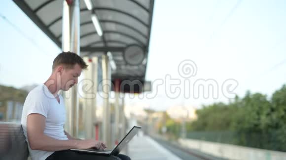 年轻英俊的观光客坐着坐着用手提电脑等火车视频的预览图