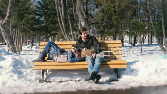 男人和女人一起在冬季城市公园的长凳上休息冬日阳光明媚男人在电话里自拍视频的预览图
