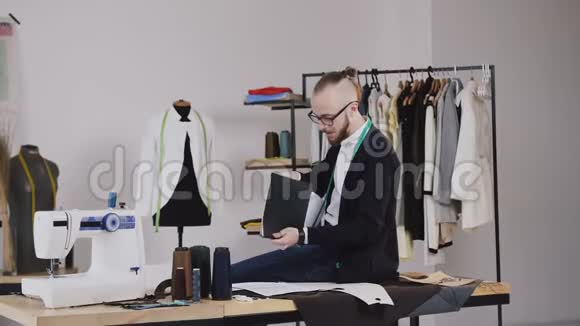 英俊的时装设计师坐在Atelier的桌子上拿着一个带有草图的文件夹他在笔记本上做笔记视频的预览图