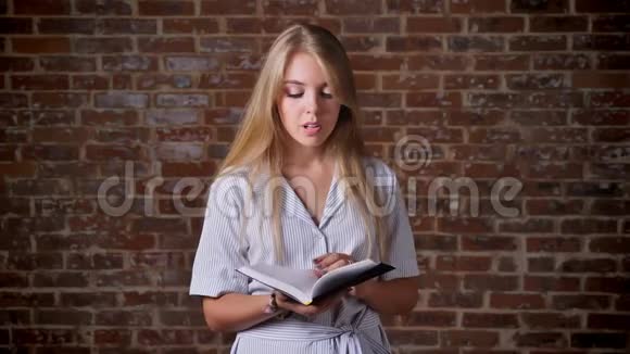 年轻的白种人金发碧眼的阅读书籍和看镜头肖像砖墙的背景视频的预览图