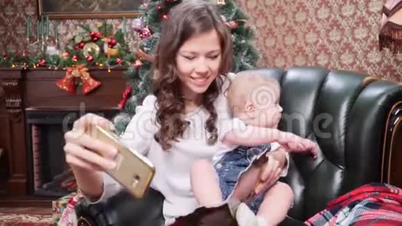 一位年轻的母亲和一个小儿子在圣诞树附近的智能手机上自拍圣诞快乐节日快乐视频的预览图