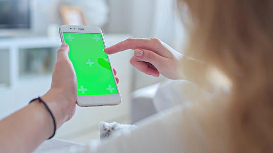 穿着白色牛仔裤的年轻女性躺在沙发上在绿色屏幕上使用智能手机视频的预览图