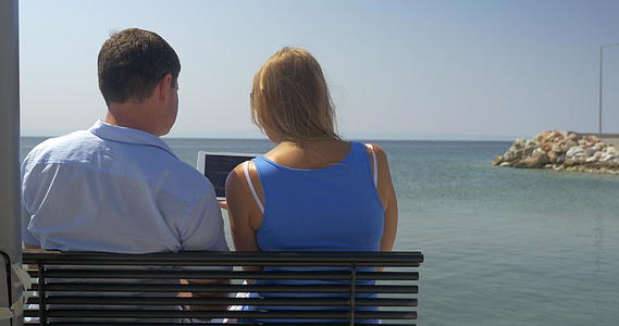 年轻的男人和女人坐在沙滩上的长椅上背景是海边的天际线看着平板长椅视频的预览图