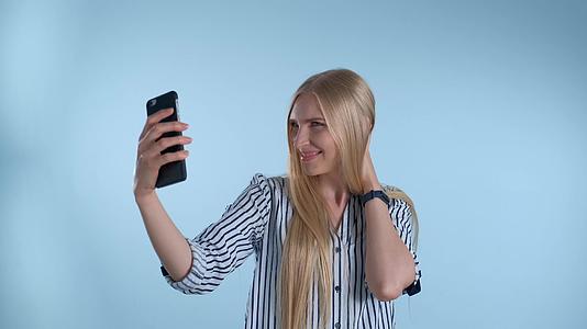迷人的金发美女在蓝色背景的智能手机上自拍视频的预览图