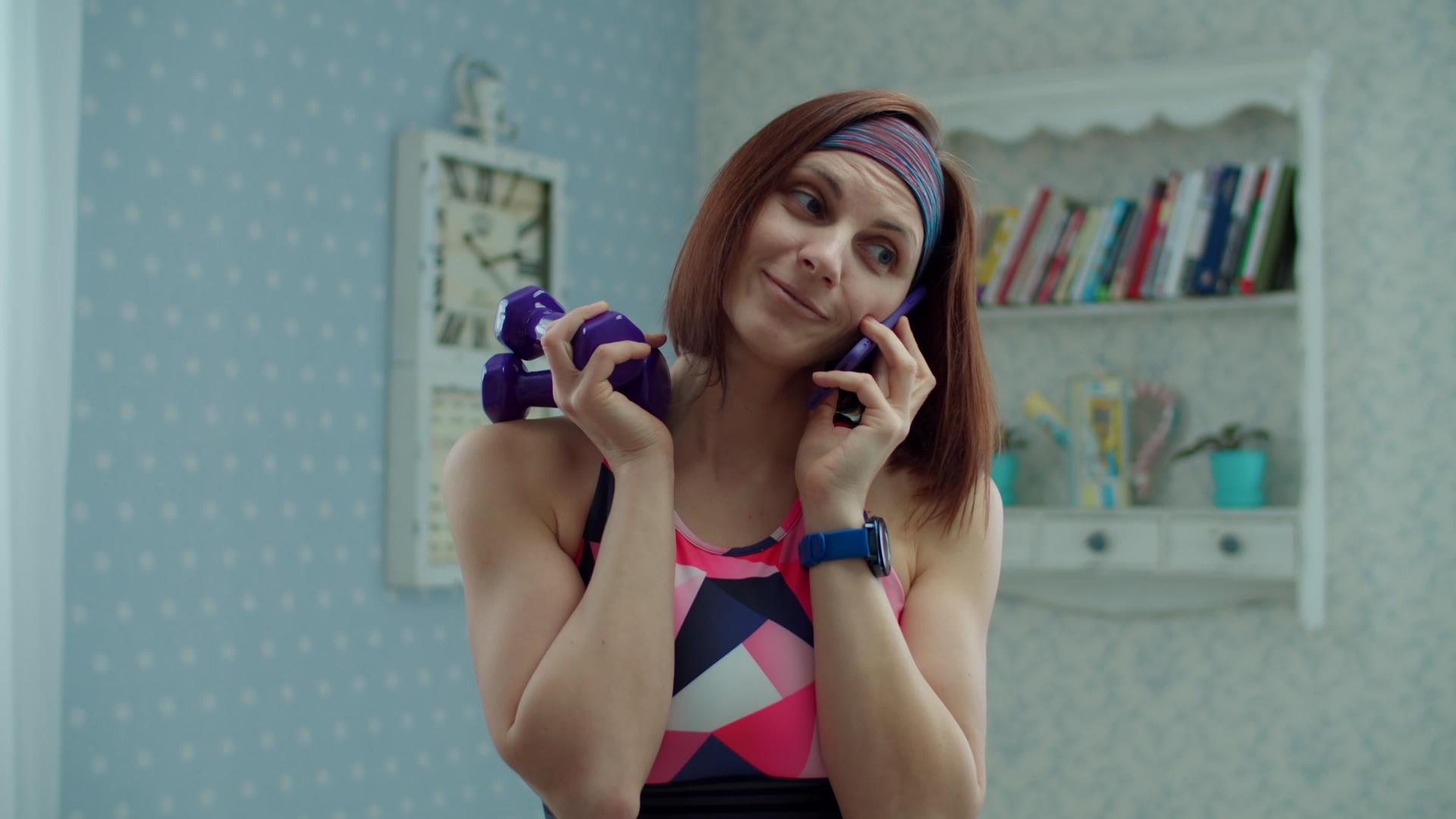 一名30岁的年轻女子穿着运动服手里拿着哑铃和手机在家里打电话视频的预览图