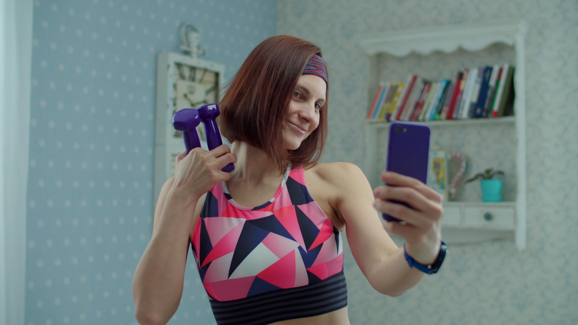 这位30岁的年轻女子穿着运动服手里拿着哑铃用手机拍着自拍视频的预览图
