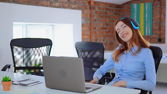 快乐的年轻女子用笔记本电脑和耳机坐在办公桌前视频的预览图