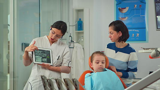 儿科牙医在平板电脑屏幕上向母亲展示X光牙齿视频的预览图