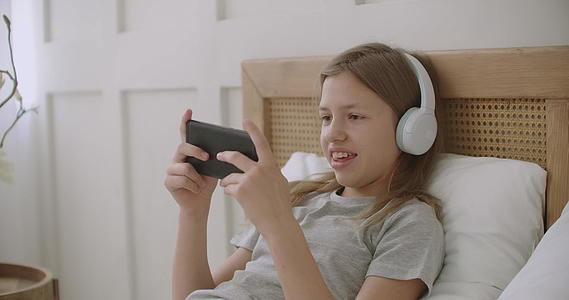 快乐的小女孩手里拿着智能手机躺在床上在网上玩游戏视频的预览图