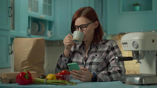 戴眼镜的年轻成年女性喝咖啡用手机站在家里的蓝色厨房里视频的预览图