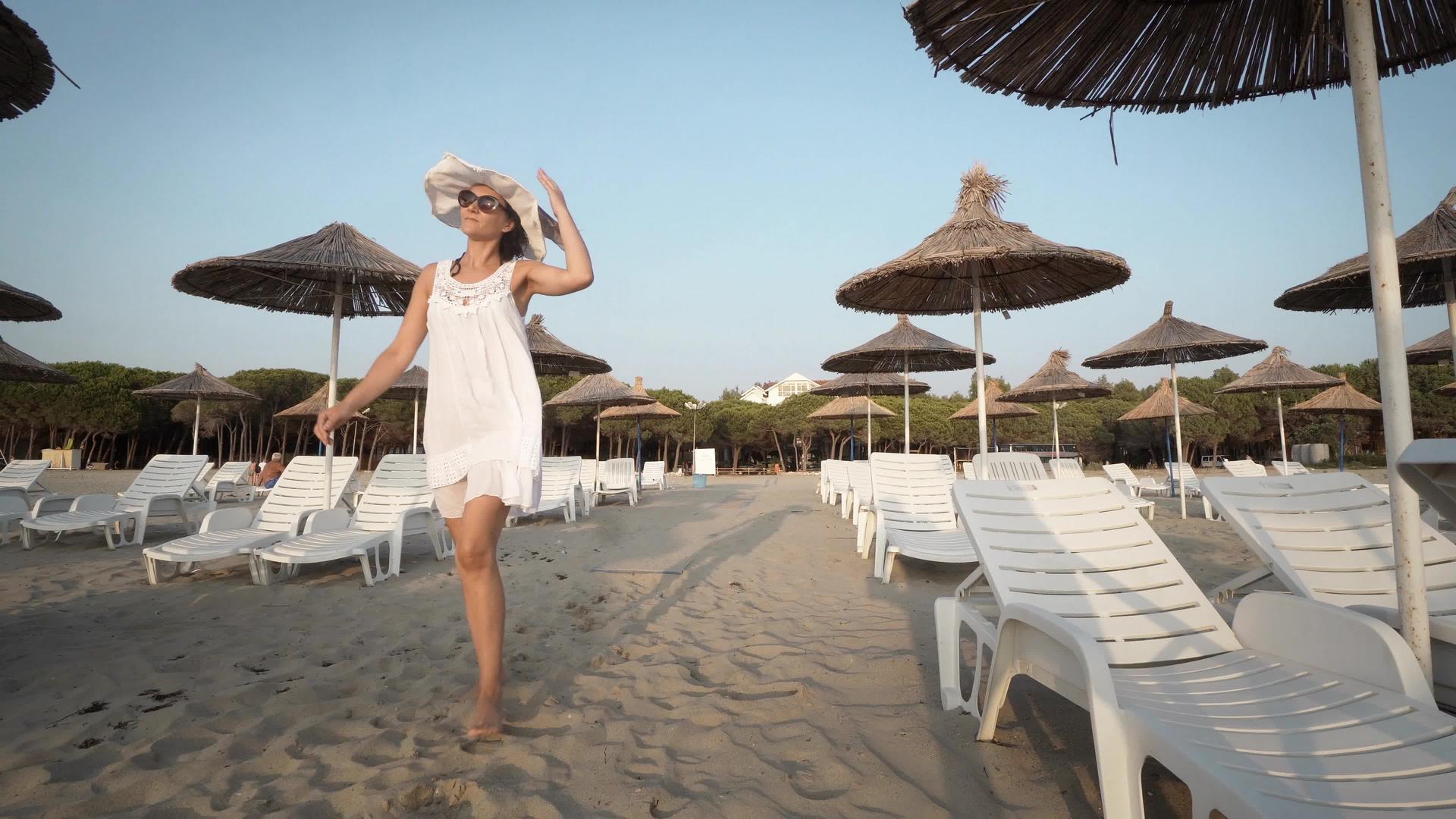 穿着白色裙子和帽子的性感女性在空旷美丽的海滩上行走视频的预览图