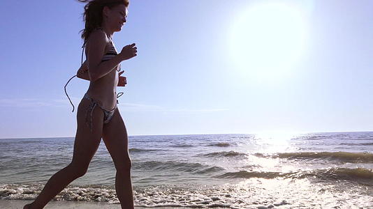 快乐美丽的女孩穿着比基尼日落时在海滩上奔跑微笑着视频的预览图
