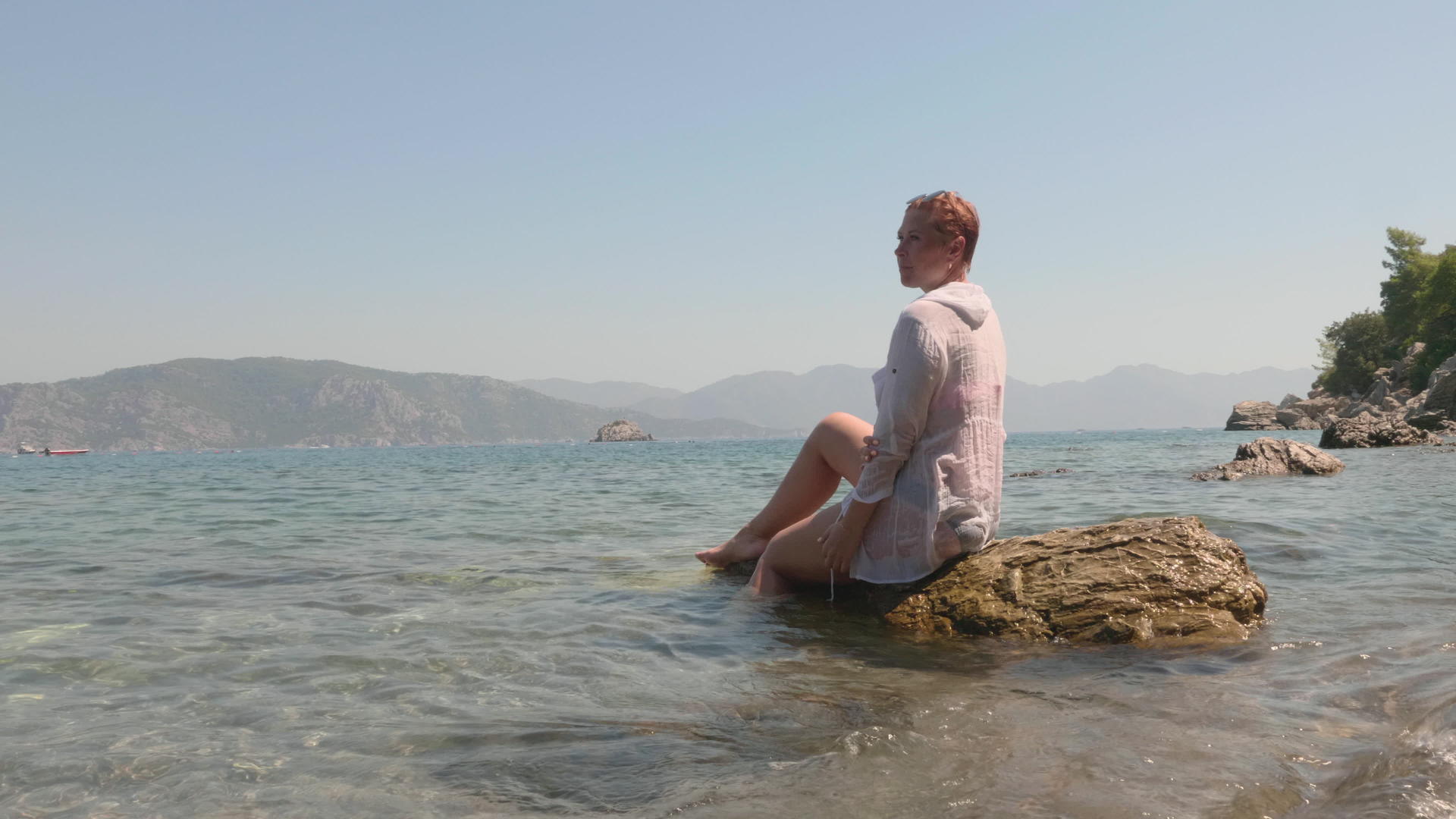 一个穿着白色湿腰外套的放松女人坐在波浪的石头上视频的预览图