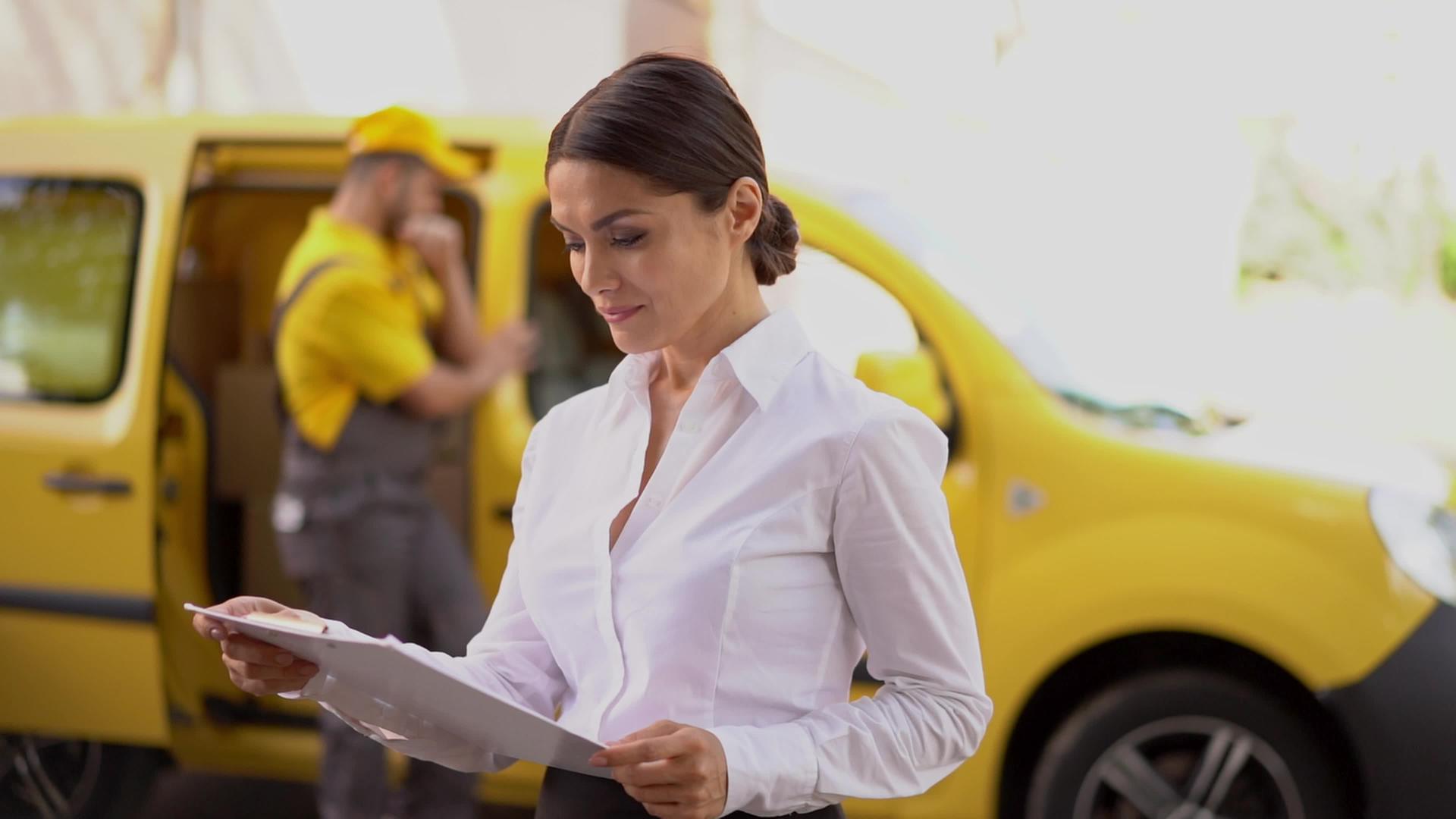 微笑的女孩在剪贴板上看到了黄色汽车附近的送货文件的信使视频的预览图