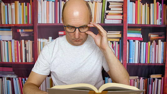 在家庭办公桌旁用眼镜看书的男士有书架背景视频的预览图