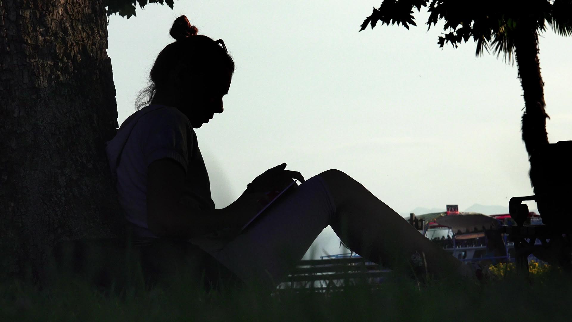 在公园里看书的年轻女子坐在树旁关上了夜幕视频的预览图