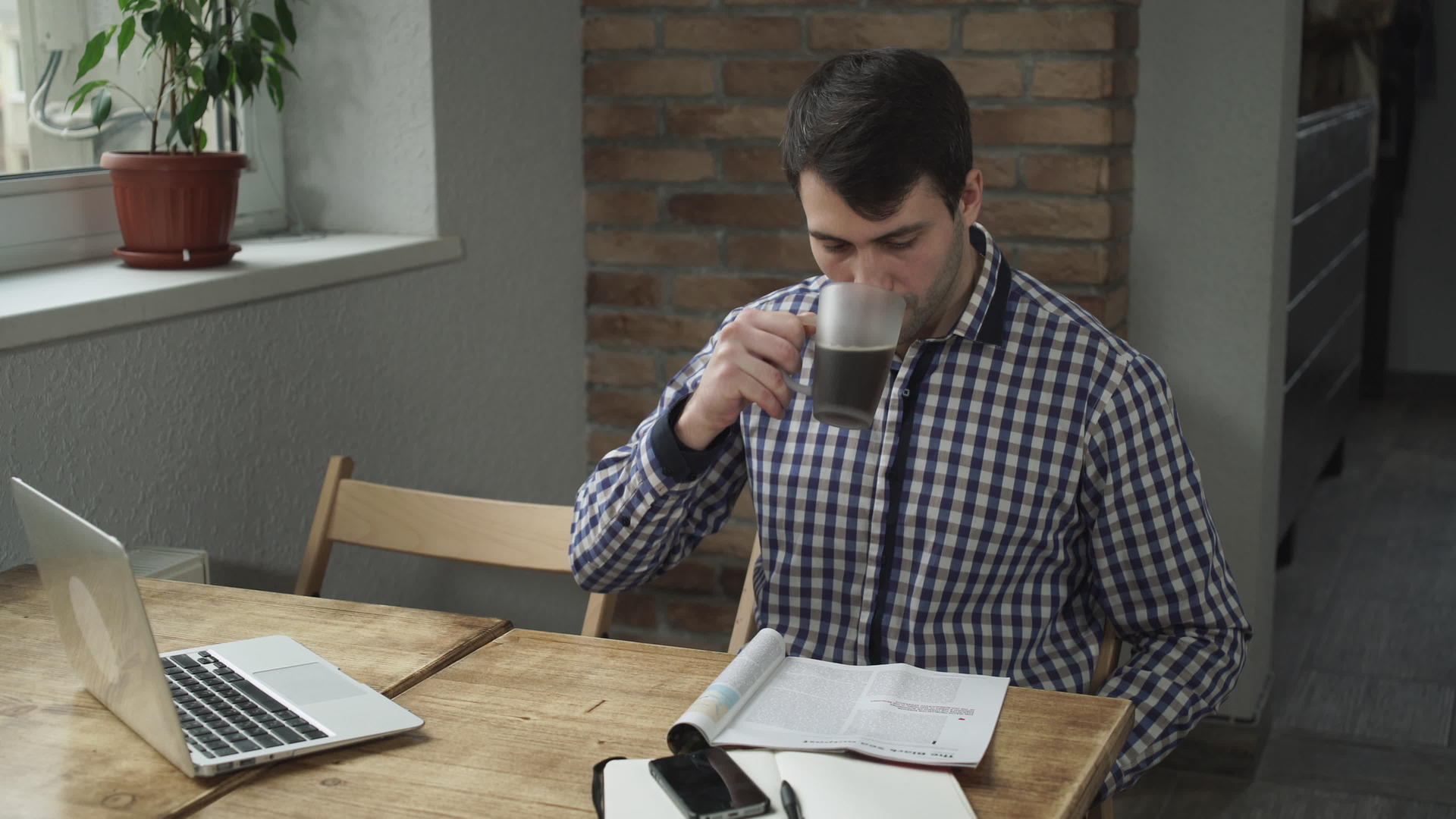 吸引人的男人喝咖啡在桌子上看报纸电话和笔记本电脑视频的预览图