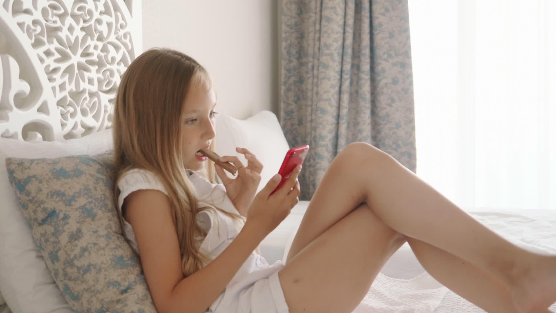 十几岁的女孩在白色的卧室里吃巧克力看着漂亮的智能手机视频的预览图