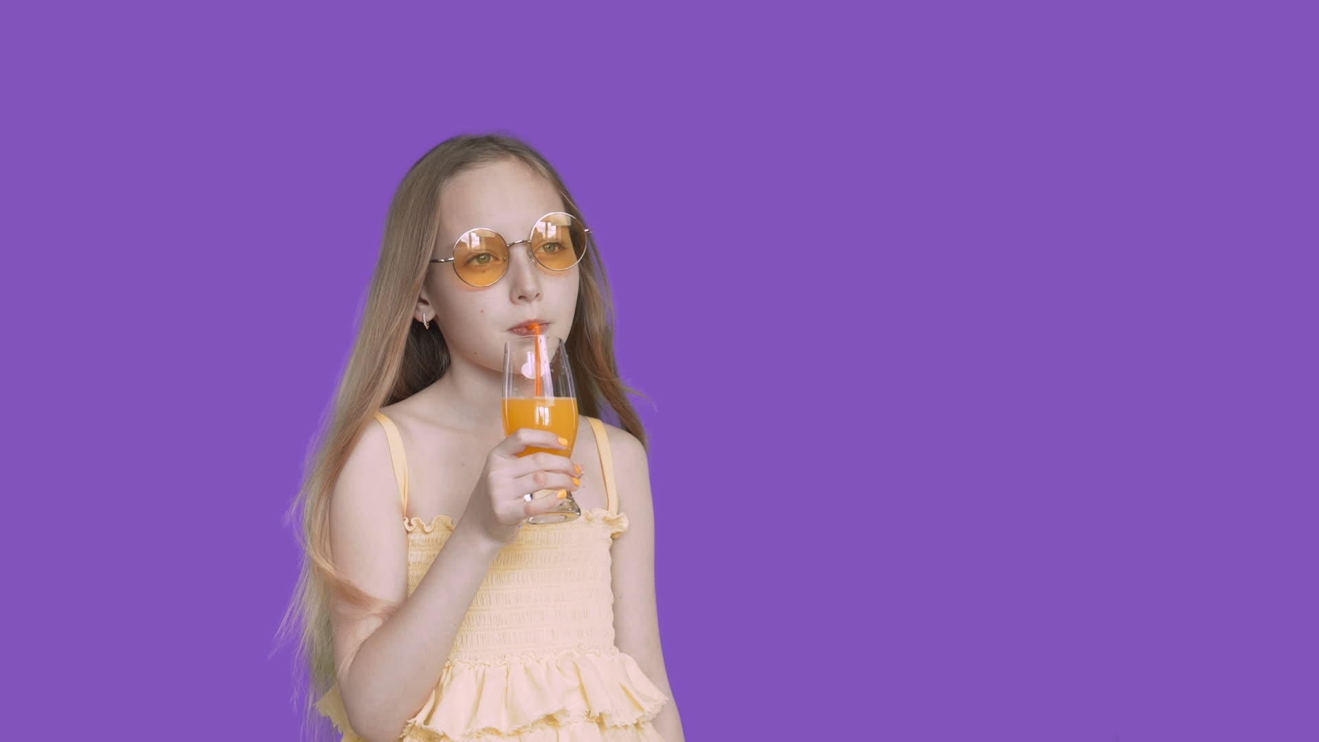 女孩在紫色背景上用稻草从玻璃杯里喝橙汁戴上黄色的黄色视频的预览图