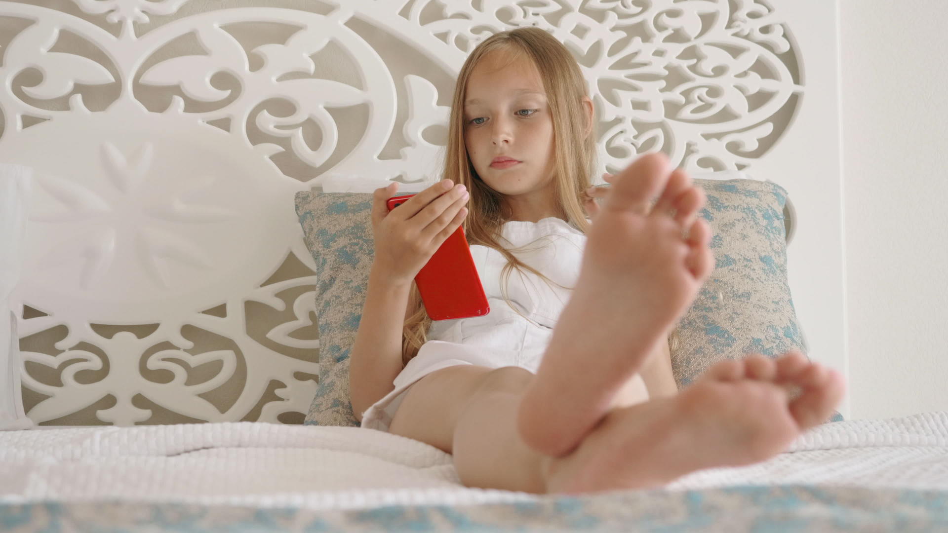 十几岁的女孩在卧室的白色床上看着智能手机赤脚的年轻女孩视频的预览图