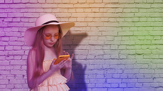 在五颜六色的灯光下十几岁的女孩在砖墙背景上浏览手机视频的预览图