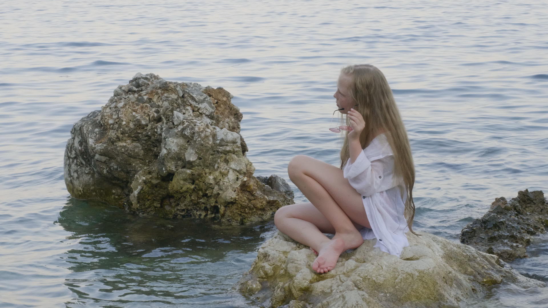 一个穿着白色束腰外衣的女孩坐在夏天海滩上的岩石上视频的预览图