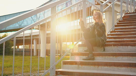 一个穿着黑色皮夹克的女孩坐在阳光背景的楼梯上视频的预览图