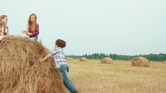 十几岁的男孩在以干草堆为背景的农田上奔跑女孩们坐在村子里视频的预览图