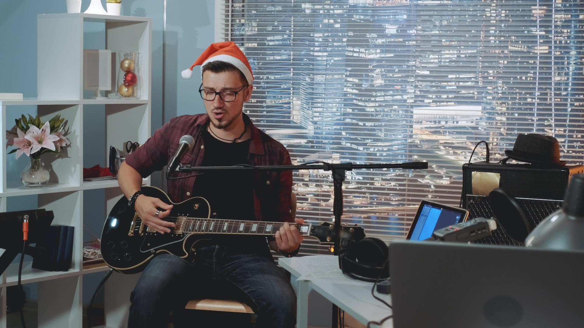 家庭录音室的年轻圣塔帽歌手在麦克风和吉他中唱圣诞歌曲视频的预览图