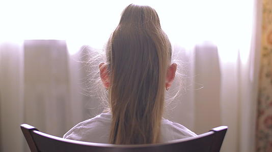 年轻的金发女孩的头发和尾巴坐在家庭房间的椅子前窗上视频的预览图