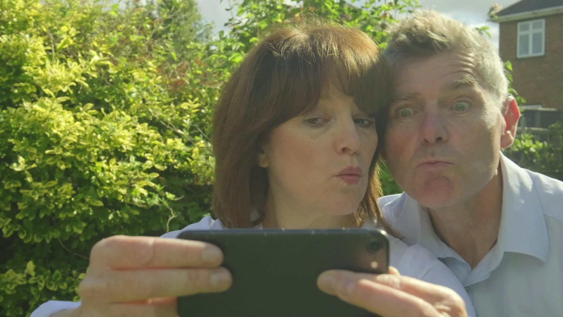 高加索夫妇在花园里的智能手机上自拍和做鬼脸视频的预览图