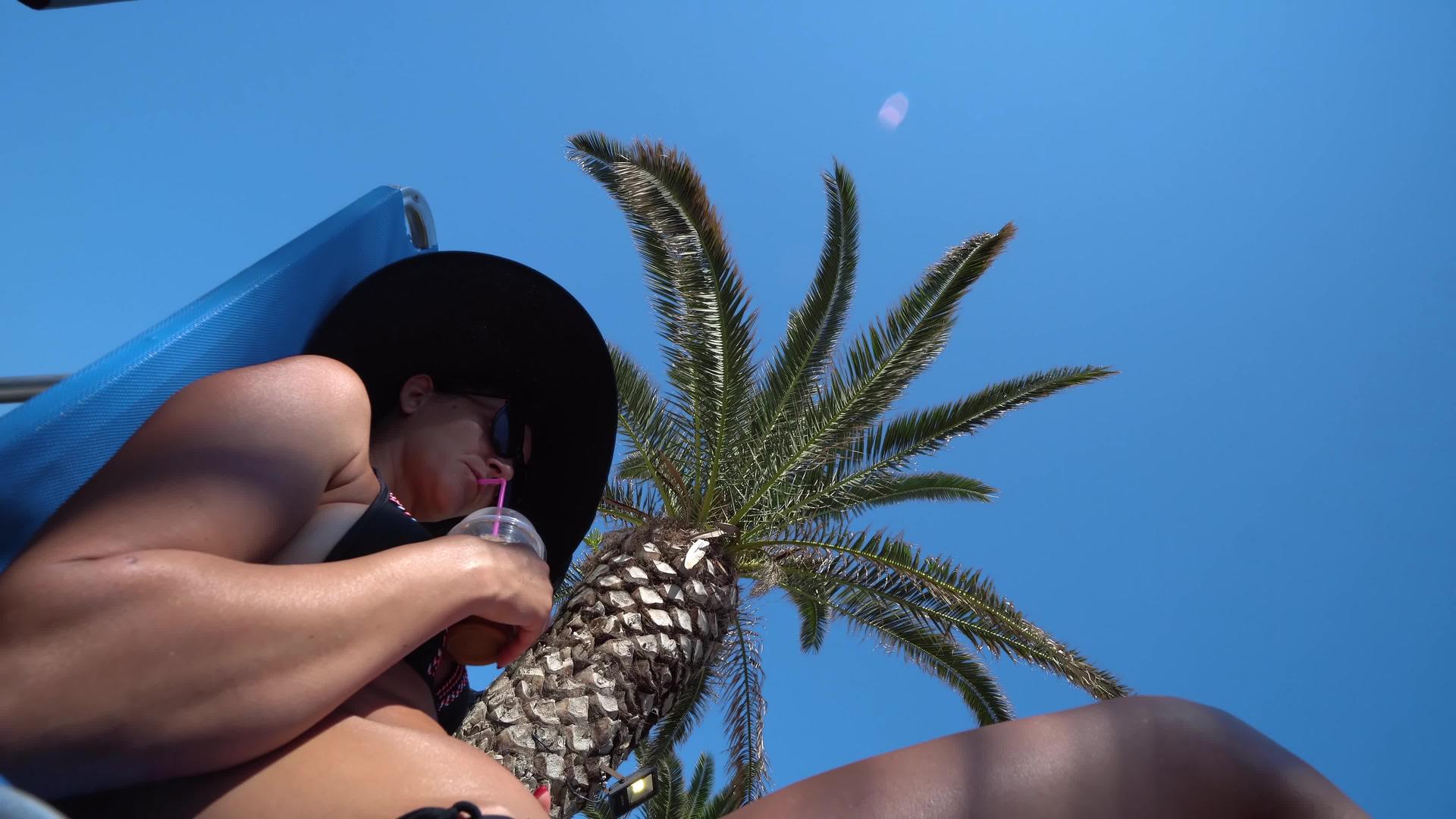 女人在棕榈树下的海滩上放松喝鸡尾酒视频的预览图