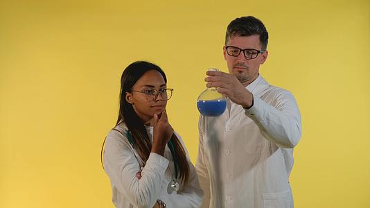 多民族男女穿着实验室外套在黄色背景下使用实验液体视频的预览图