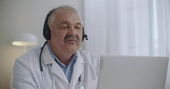 年长的医生穿着白色长袍脖子上有听诊器正在用笔记本电脑录像视频的预览图