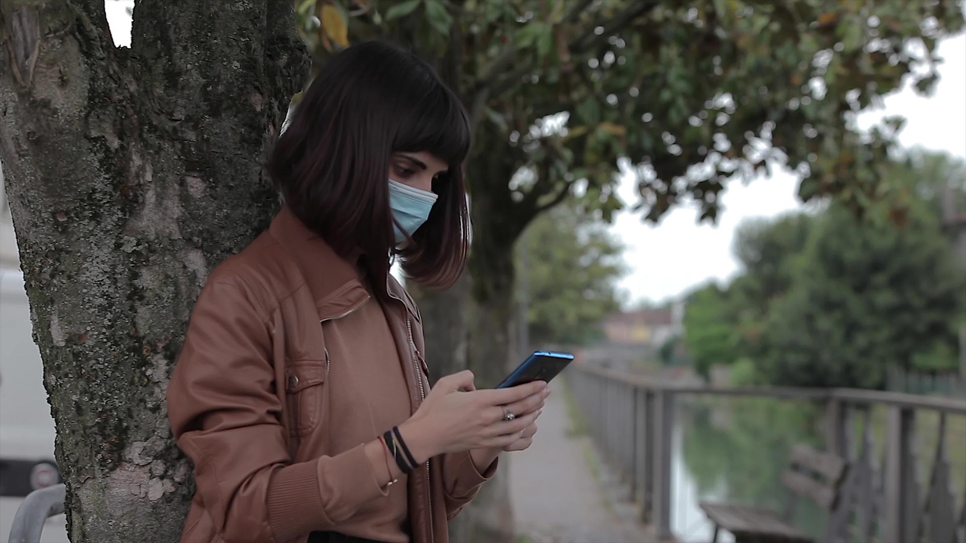 在2号公园戴着医用口罩的年轻女孩看着她的智能手机视频的预览图