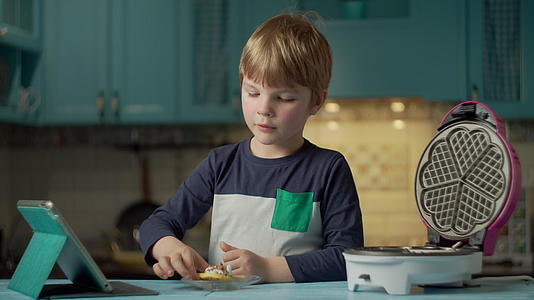 在看平板电脑时男孩用奶油和巧克力吃自制的华夫饼干视频的预览图