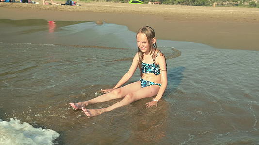 可爱的女孩穿着泳衣坐在沙滩上享受海浪视频的预览图