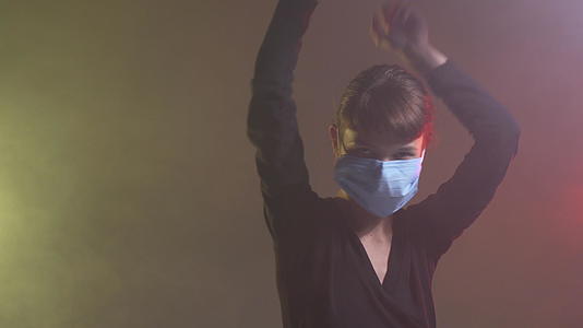 可爱的女孩戴着医用面具微笑在多彩的烟雾背景下跳舞视频的预览图