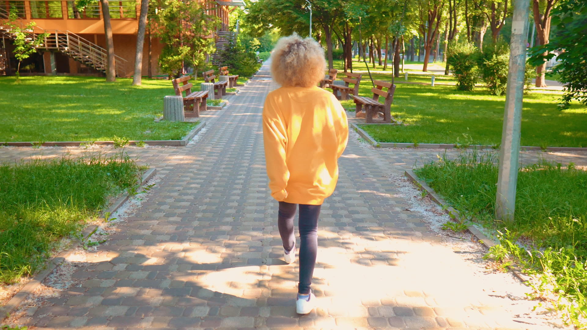 女孩留着金发和卷发夏天走在绿树成荫的街道上视频的预览图