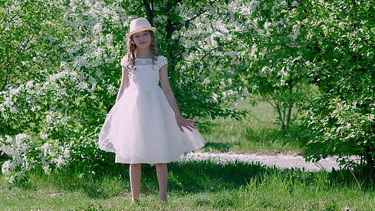这个迷人的女孩穿着白色的夏装戴着帽子在果园里盛开的果树旁边视频的预览图