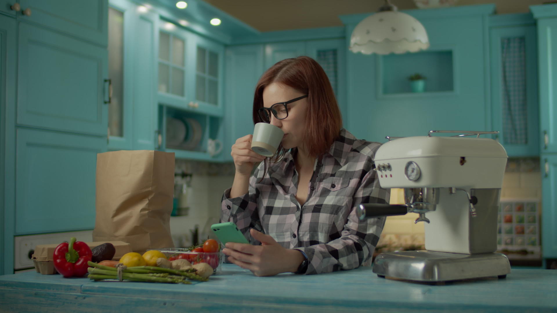 戴眼镜的年轻成年女性喝咖啡用手机站在家里的蓝色厨房里视频的预览图