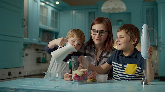 快乐的母亲和两个孩子在家里的蓝色厨房里用搅拌机做饭视频的预览图