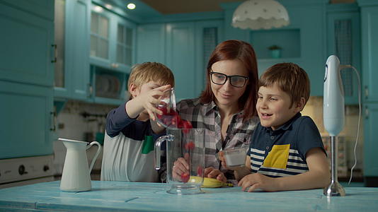 快乐的母亲和两个孩子在家里的蓝色厨房里用搅拌机做果酱视频的预览图